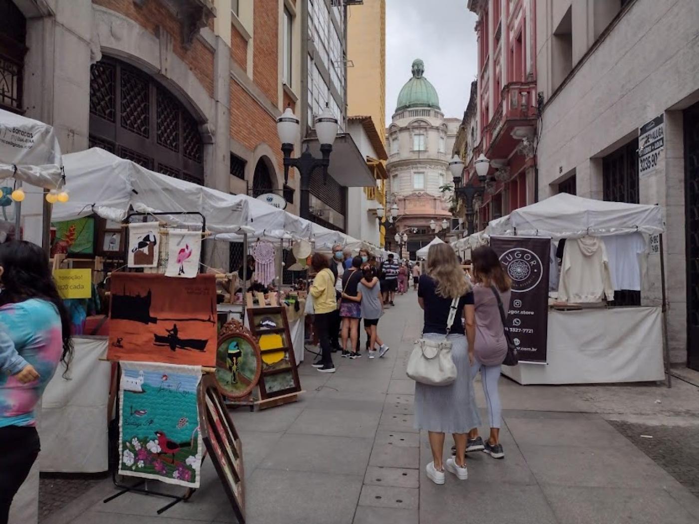 Quadros e outros produtos artesanais em barracas de boulevard, com a Bolsa do Café ao fundo. #pracegover