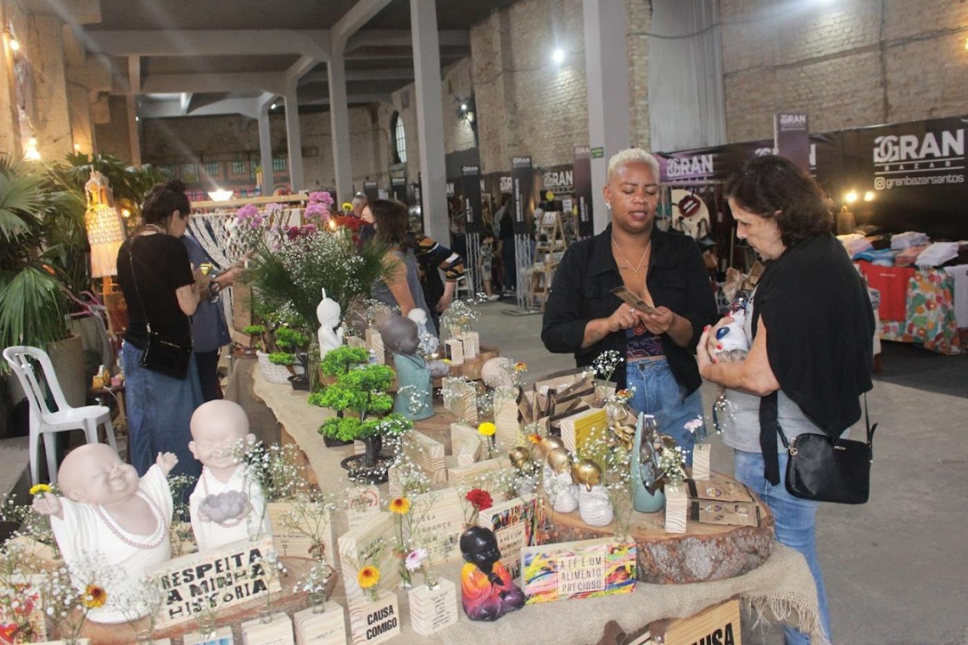 Empreendedora apresenta produto artesanal para cliente em feira. #pratodosverem