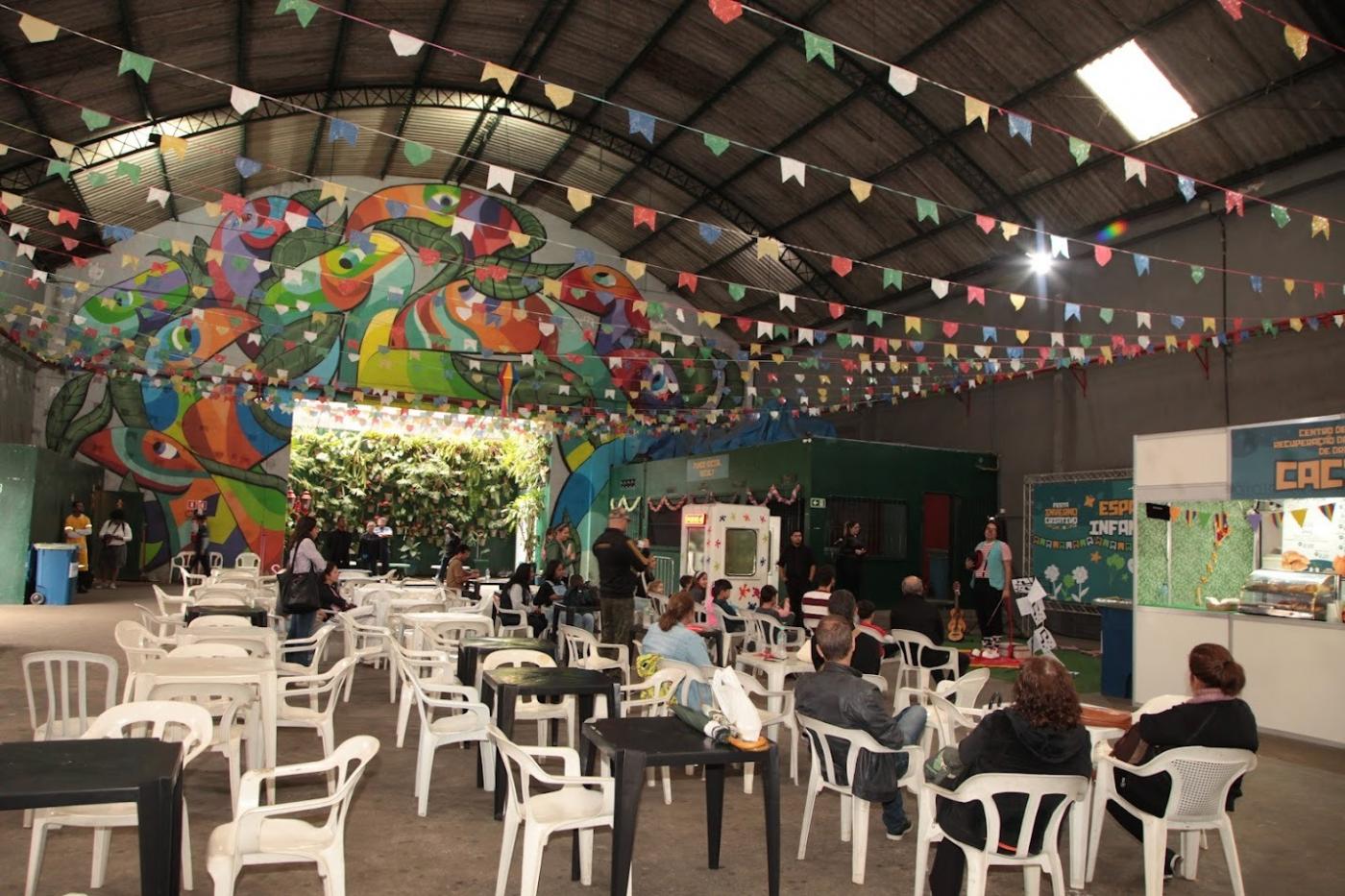 galpão decorado com bandeirinhas, mesas e cadeiras dispostas com algumas pessoas sentada em torno