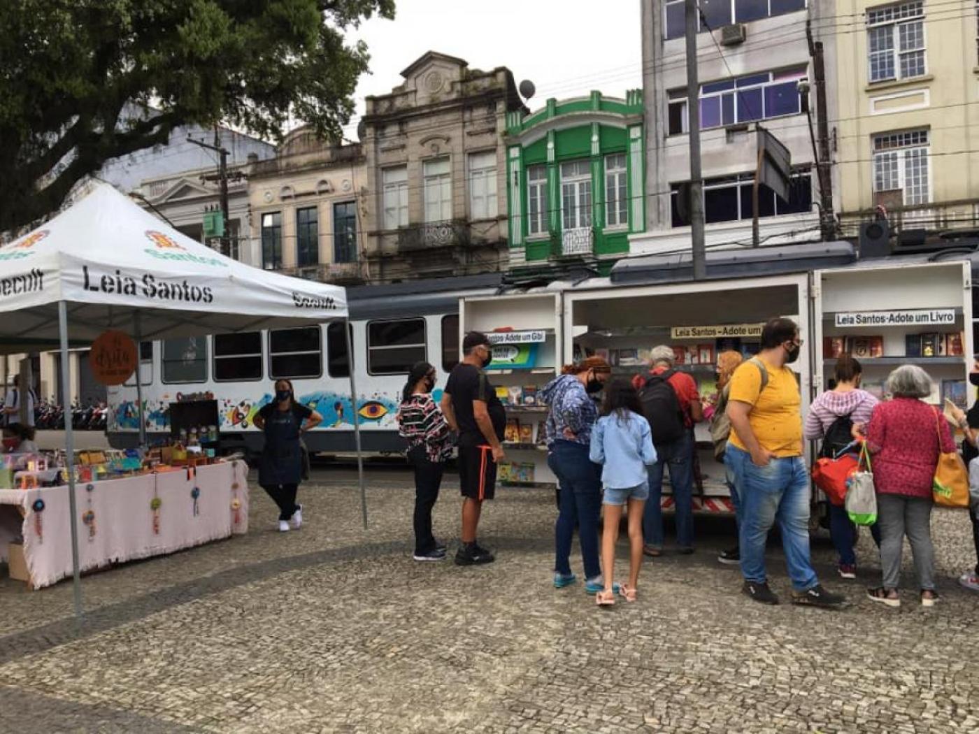 Pessoas na praça olhando estandes com livros #paratodosverem