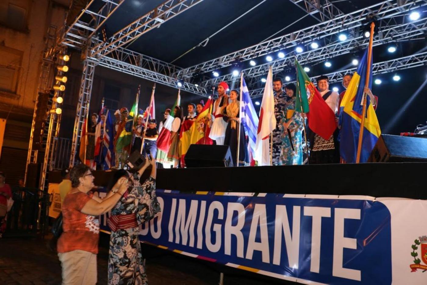 Pessoas com bandeiras de diversos países em cima de palco #paratodosverem