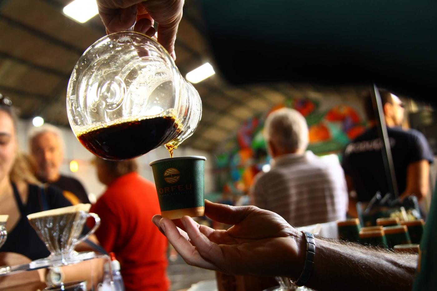 imagem em close mostra mão segurando bule e depositando café em um copo. Ao fundo há pessoas circulando. #paratodosverem