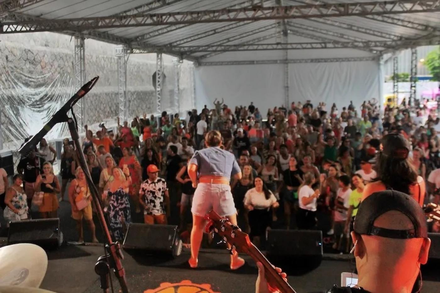 cantor se apresenta em palco com público à frente, Todos estão sob uma grande tenda. #paratodosverem 
