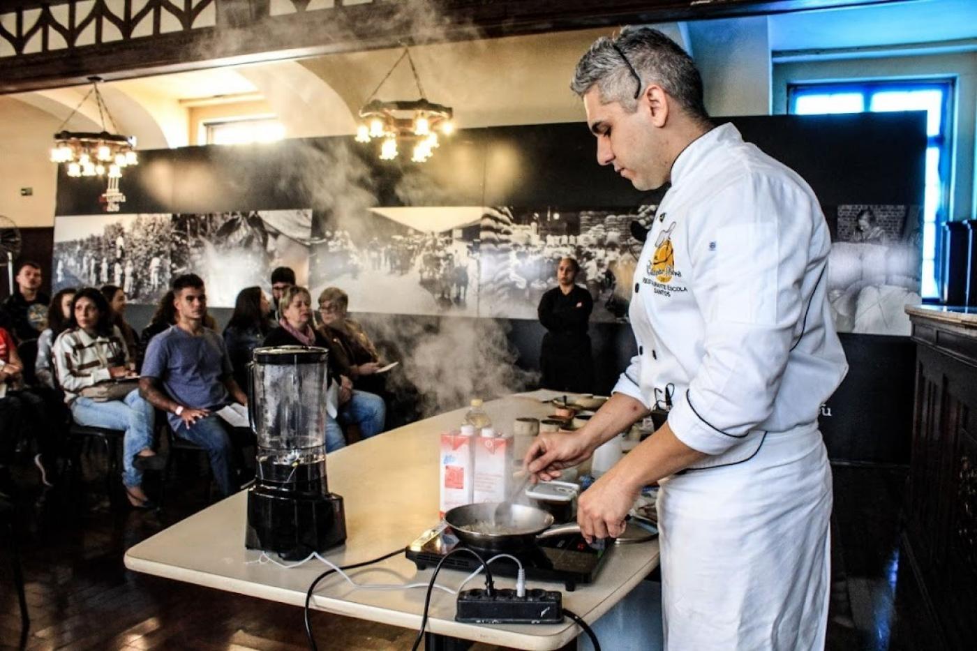 chef de cozinha está atrás de mesa preparando prato. Dá pra ver fumaça saindo da panela. Na mesa, há um liquidificador. Ao fundo, pessoas assistem à aula. #paratodosverem