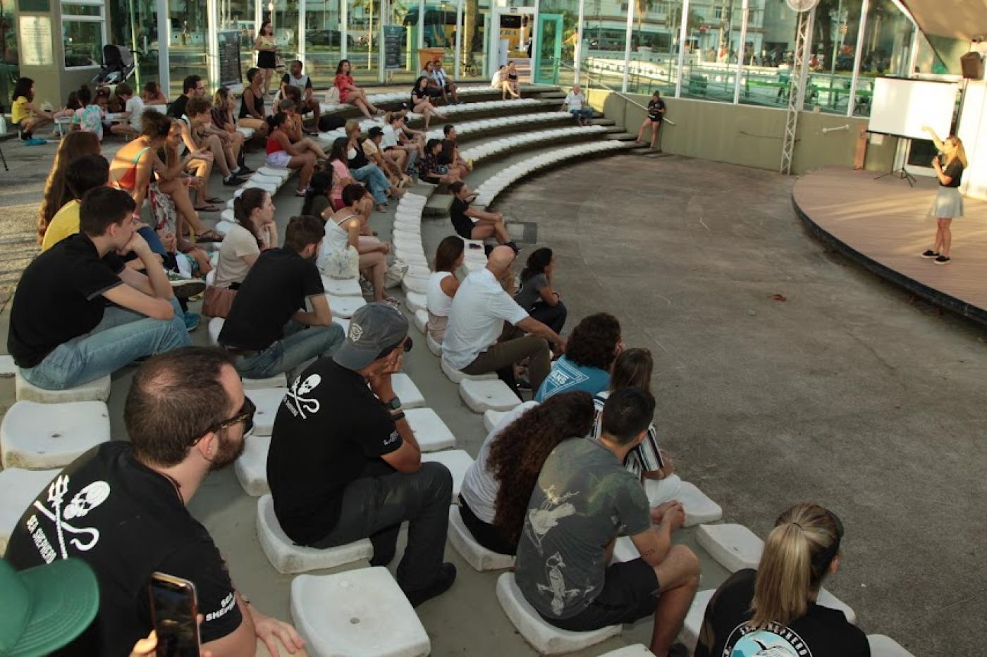 Público sentado na concha acústica assiste palestrante mulher. #pratodosverem