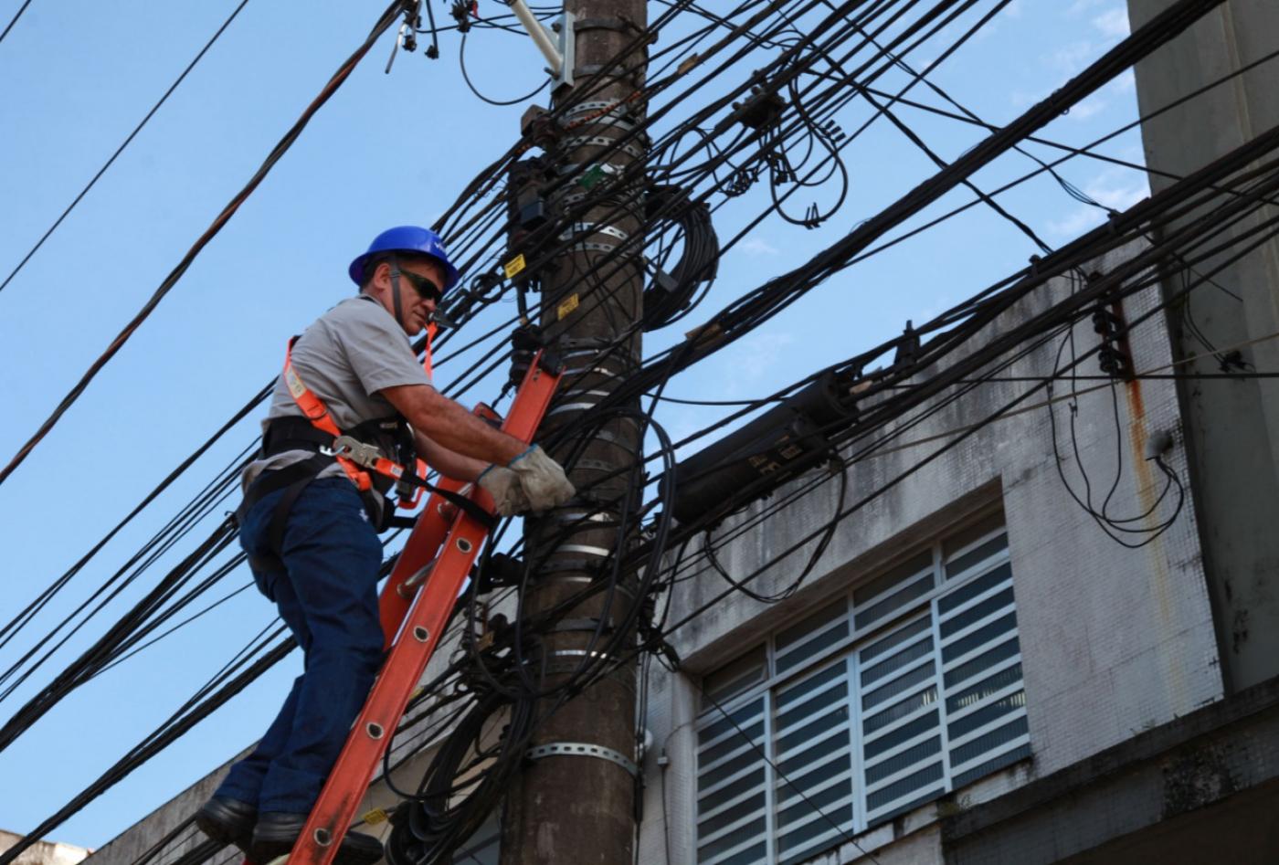 Profissional no alto de escada mexe em fiação em poste. #pracegover