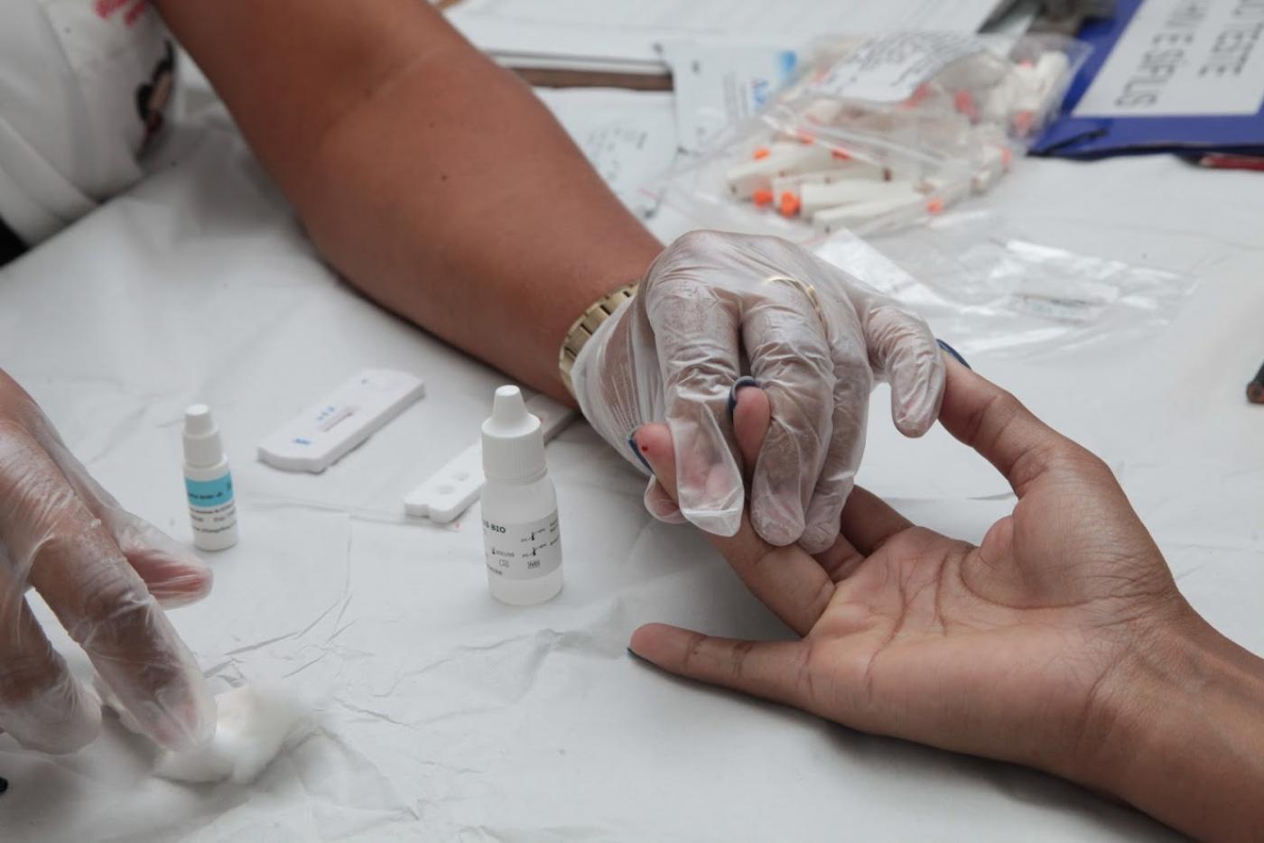 Mãos com luvas seguram a mão de alguém que acaba de fazer o teste rápido. A ponta do dedo do examinado tem uma gota de sangue. Há frascos pequenos na mesa. #Paratodosverem