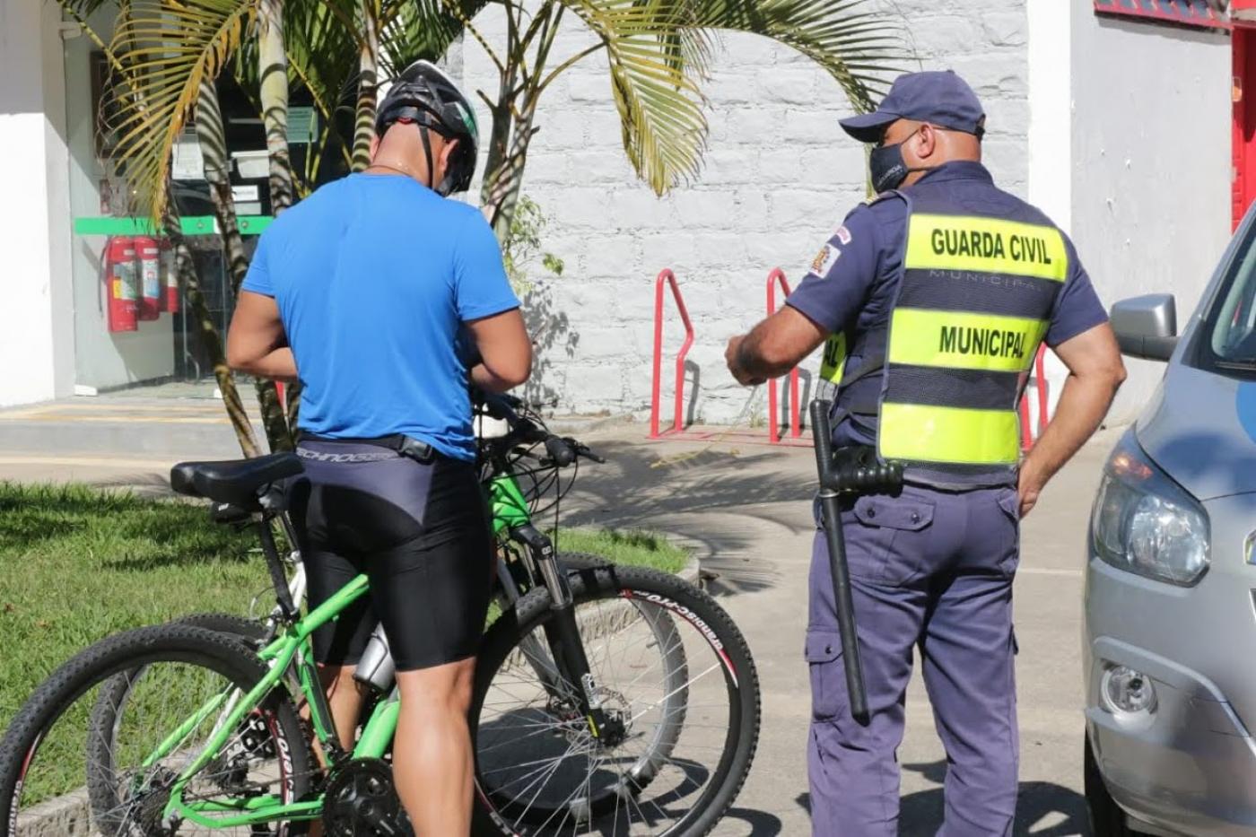Guarda municipal está ao lado de ciclista. Ambos estão de costas. #Paratodosverem