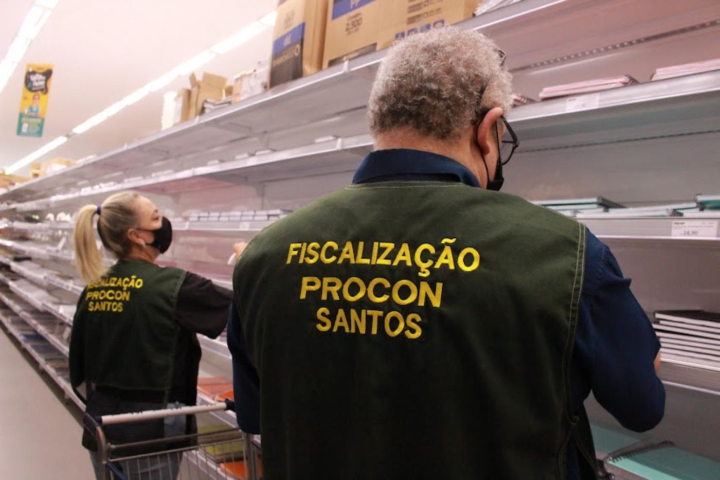 dois fiscais do procon uniformizados são fotografados de costas. Eles estão diante de prateleiras em papelaria observando produtos. #paratodosverem