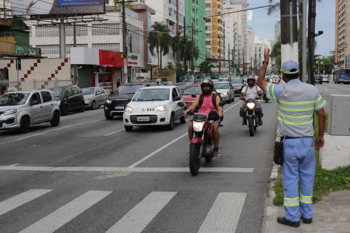 Agente da CET está no canteiro central de avenida sinalizando para motoristas. Ele está ao lado de uma faixa de pedestres. #Paratodosverem