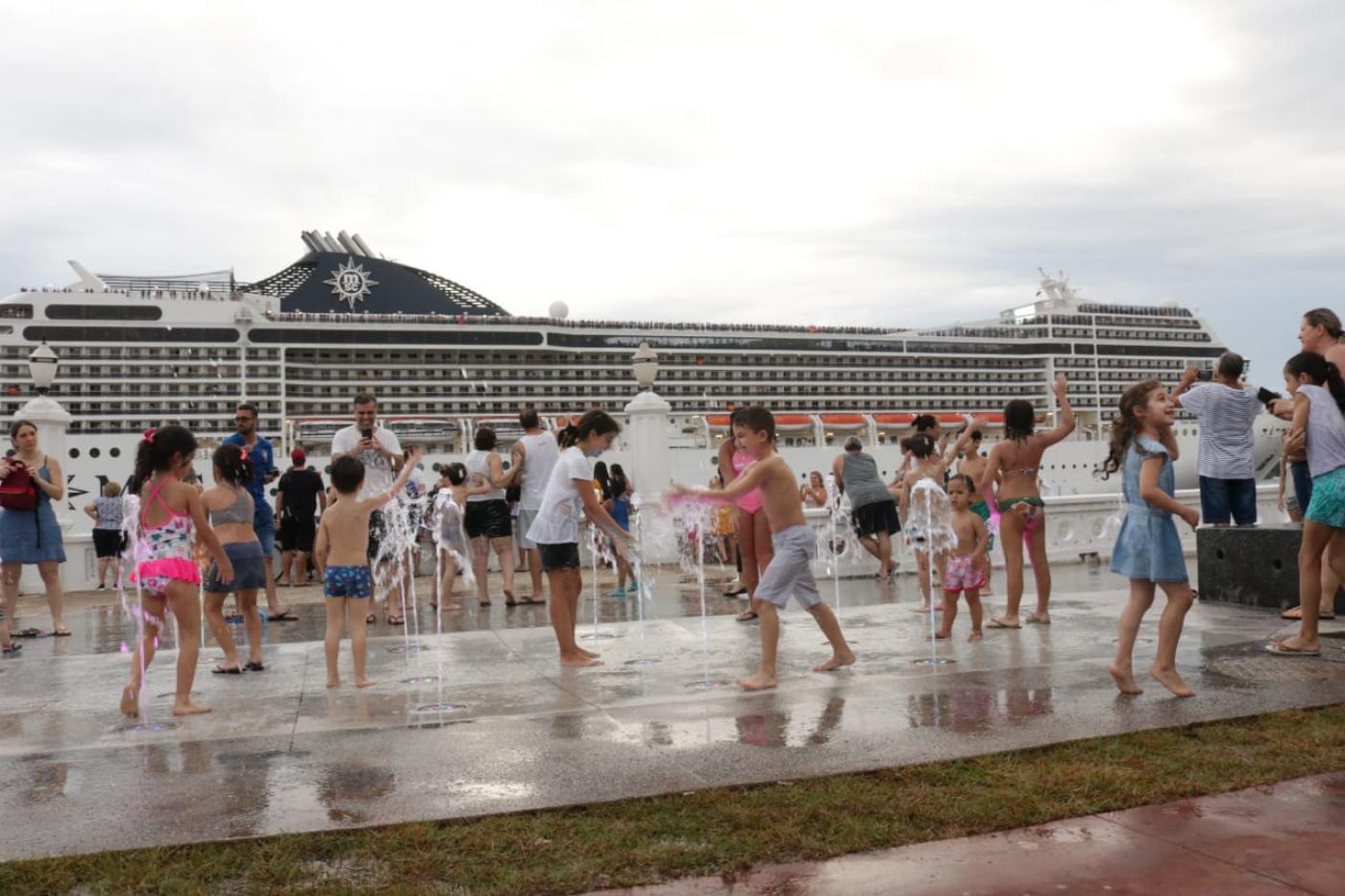 Crianças brincam como água da fonte interativa com navio de cruzeiro passando ao fundo. #pracegover