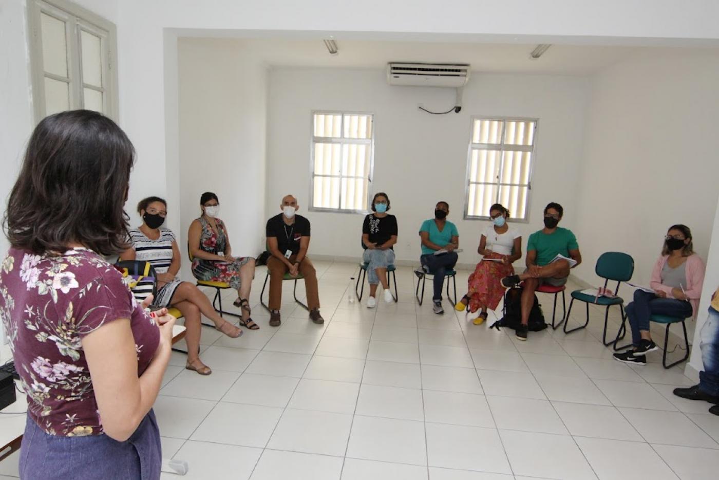 educadores estão sentados atentos a uma mulher que fala a eles. Ela está em pé, ao lado esquerdo da imagem e de costas para a foto. #paratodosverem