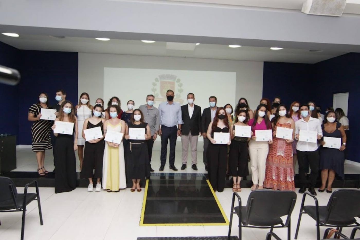 grupo de formandos posa para foto reunido em auditório. Ao fundo, o brasão de Santos está projetado num telão. #paratodosverem