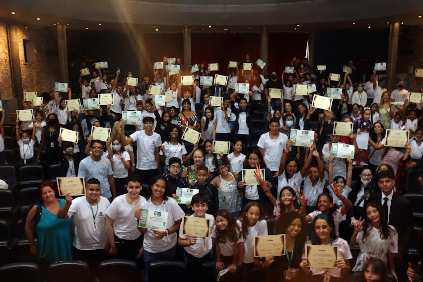 alunos estão na plateia de teatro ostentando certificados. #paratodosverem
