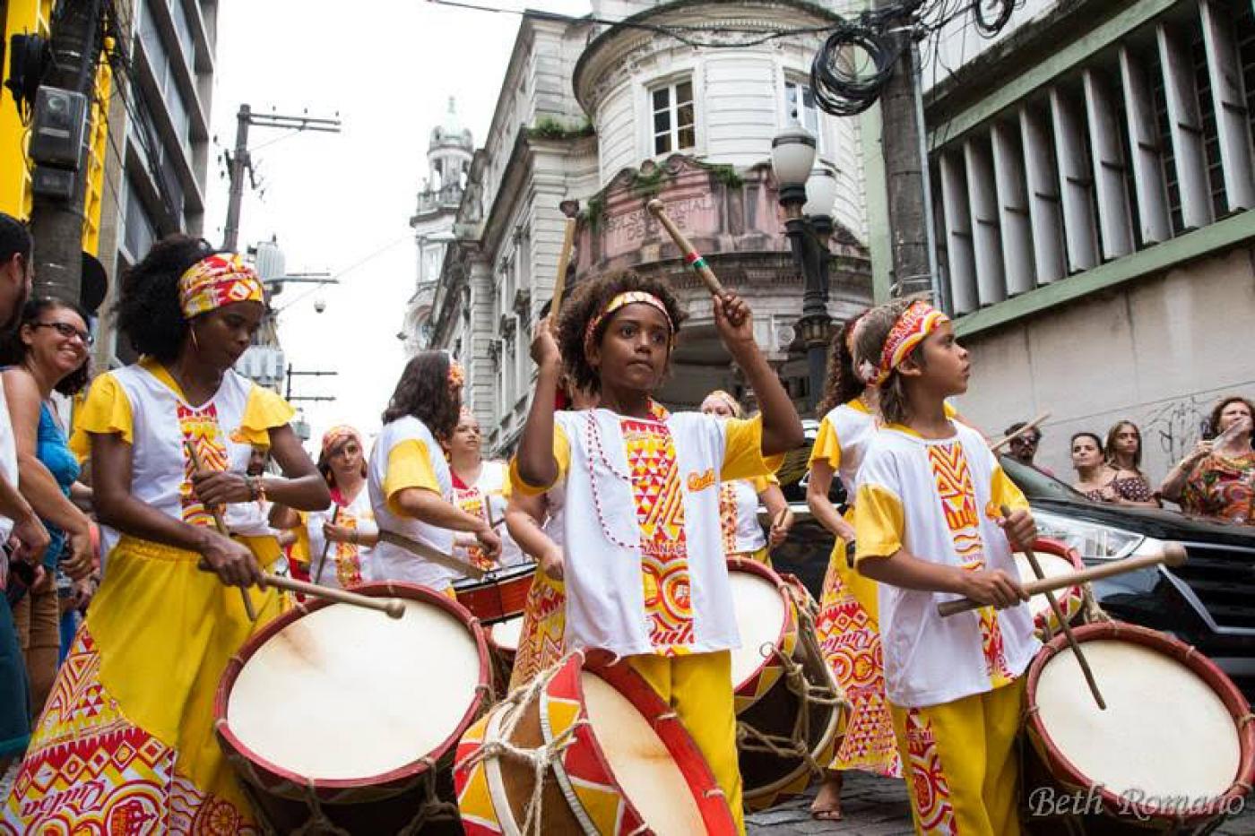 Grupo vestido com roupas do tipo afro e instrumentos de percussão desfila em rua. #Pracegover