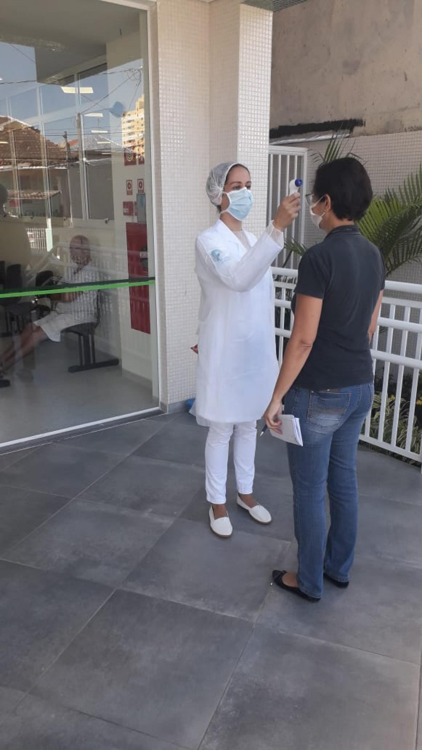 Mulher vestida de branco, usando luva, touca e máscara mede temperatura de uma mulher. Ambas estão em pé na porta de entrada de um prédio. A mulher de branco aponta um termômetro para a testa da outra. #Paratodosverem