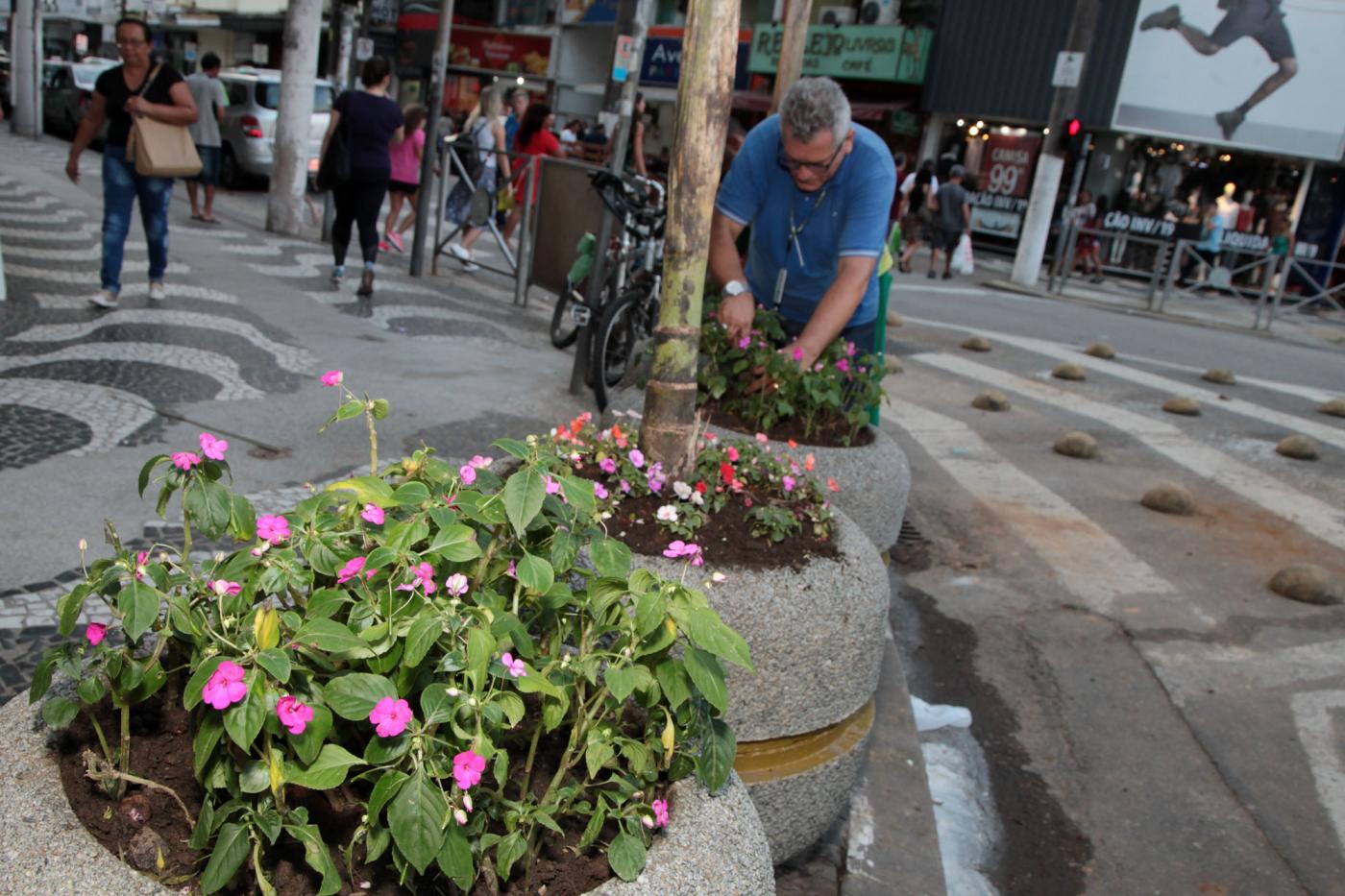 Vasos com flores em praça. Ao fundo, um funcionários planta mudas em outro vaso. #Pracegover