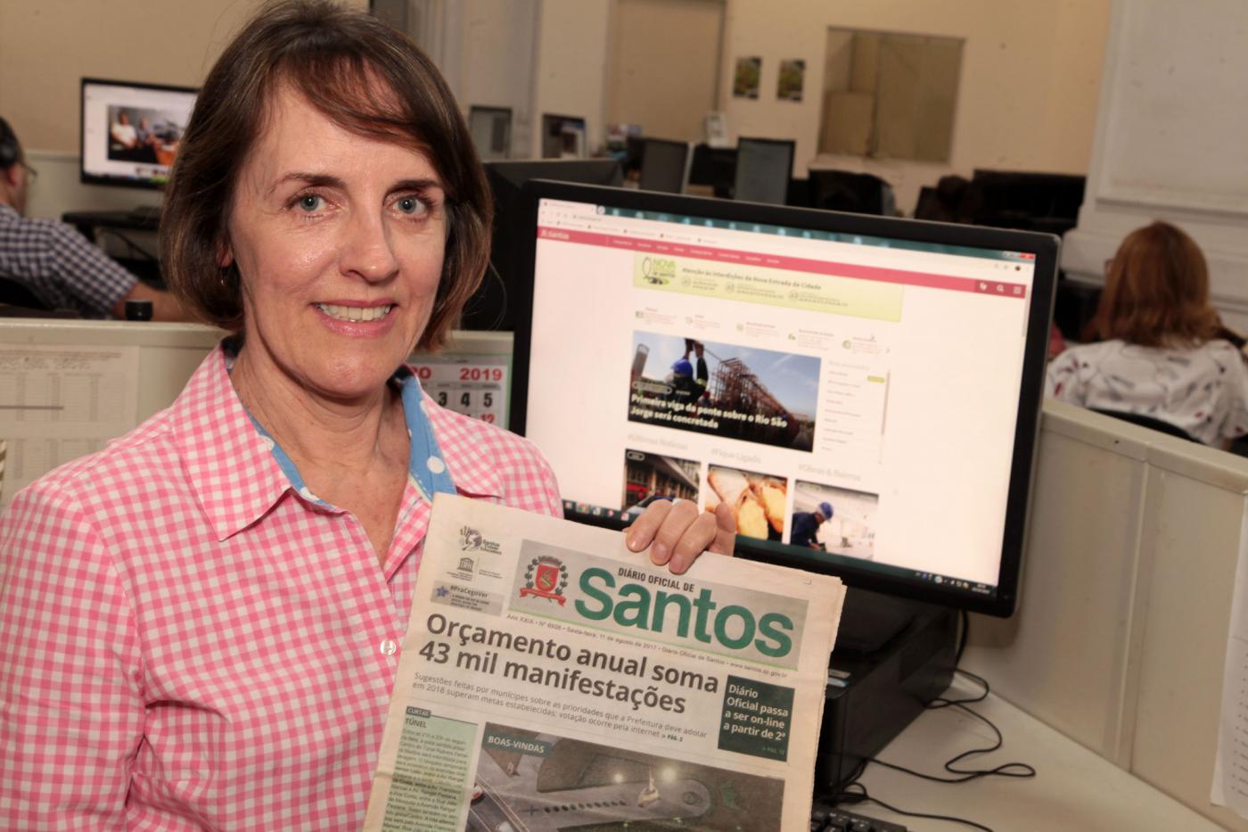 Márcia está segurando uma antiga edição do Diário Oficial em papel. Atrás dela, um monitor de desktop está aberto no  Santos Portal. #Pracegover