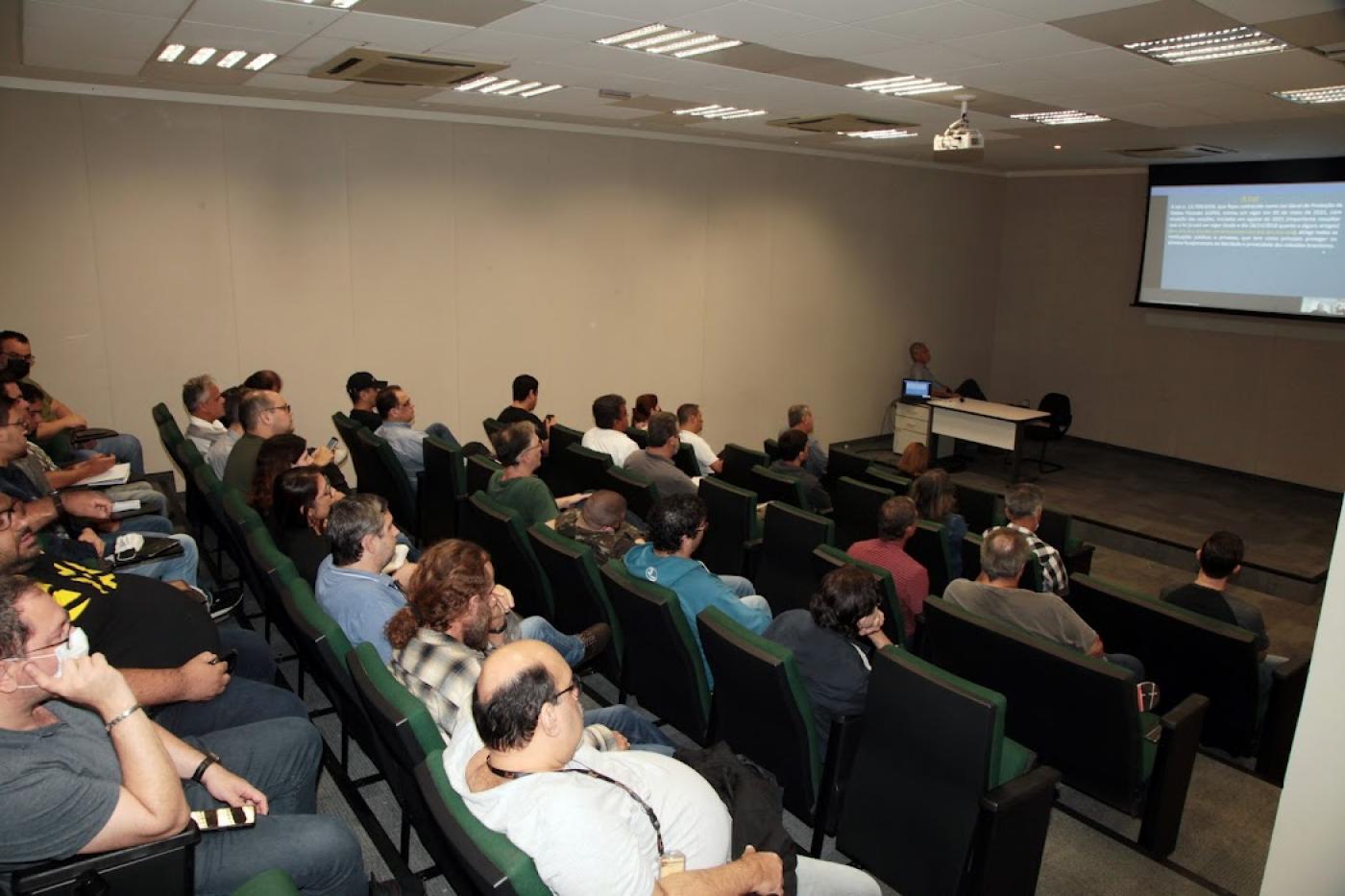 pessoas sentadas em auditório observando telão #paratodosverem