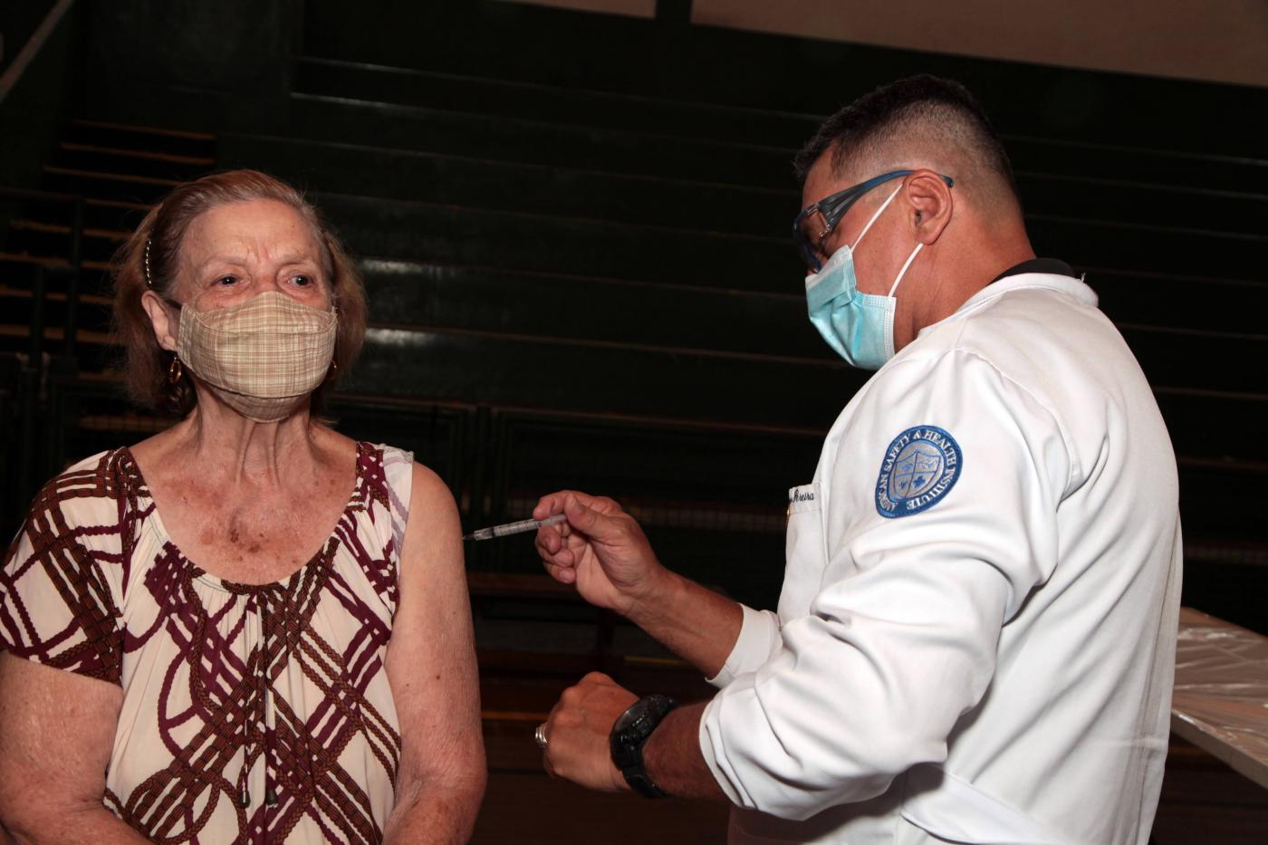 Mulher recebe vacina no braço esquerdo aplicada por uma pessoa que está de branco #paratodosverem