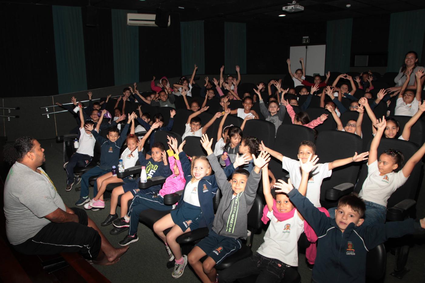 crianças estão em sala de cinema felizes, todas com os braços para o alto. #paratodosverem 