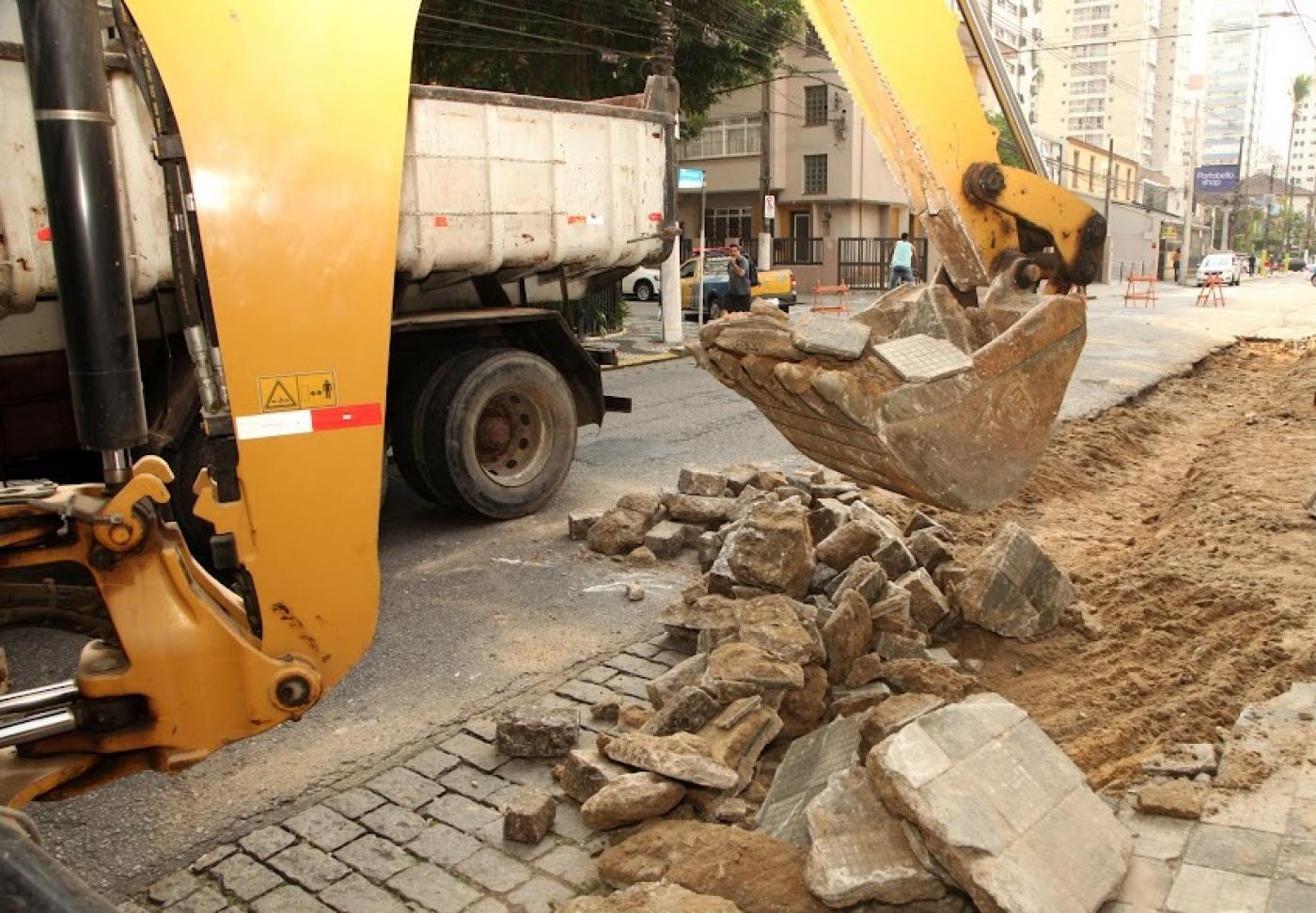 Escavadeira recolhe trecho de calçada demolido. Ao fundo se vê um desvio no trânsito. #paratodosverem