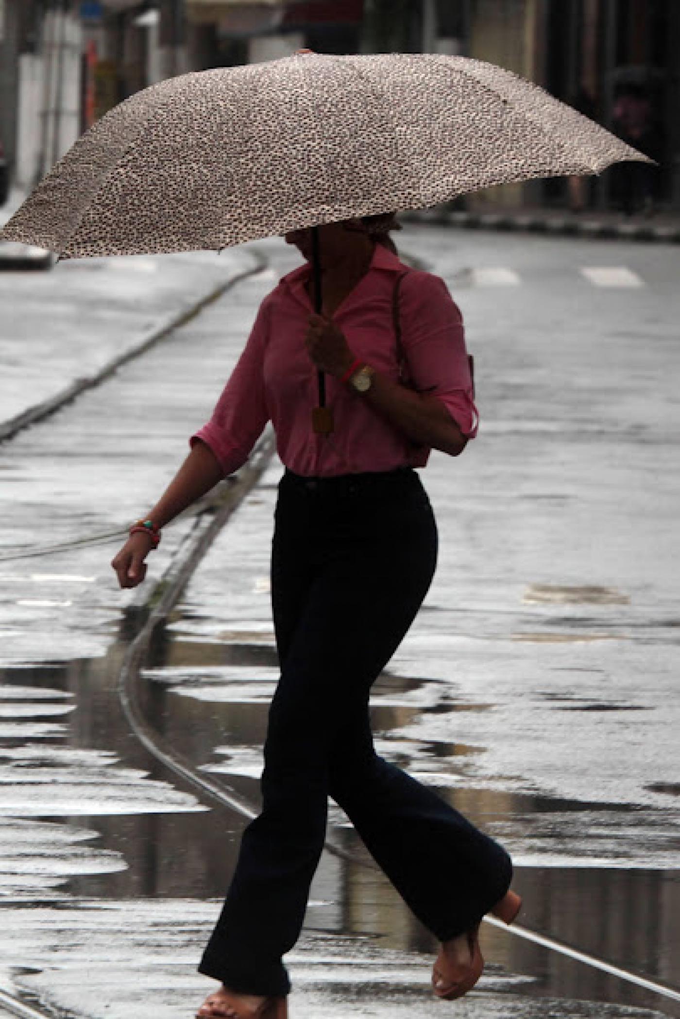 mulher atravessa rua sobre trilhos do bonde segurando guarda-chuva. Está chovendo. #paratodosverem 
