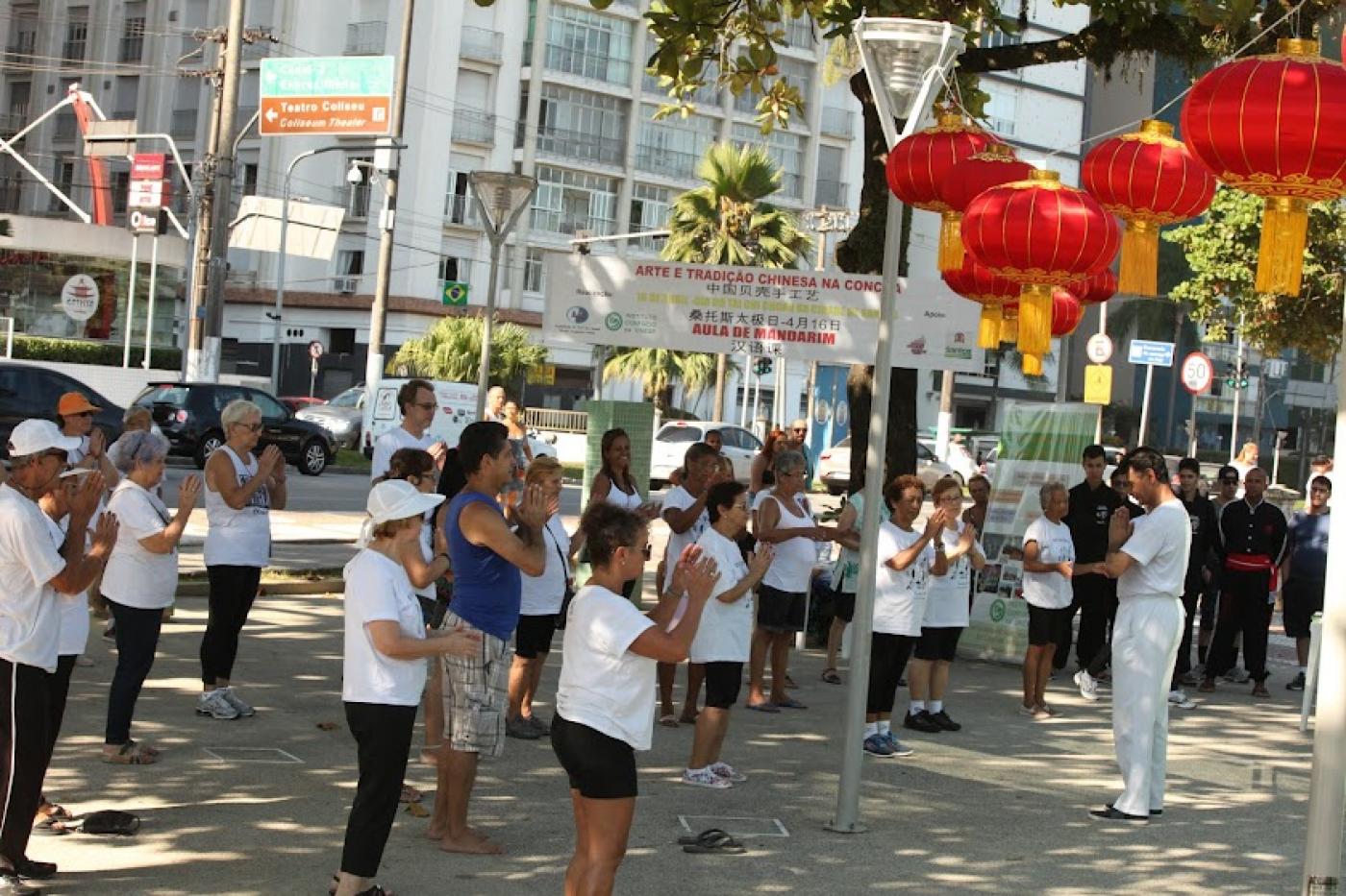 pessoas praticam tai chi chuan em praça. ambiente está decorada com luminárias orientais. #paratodosverem 