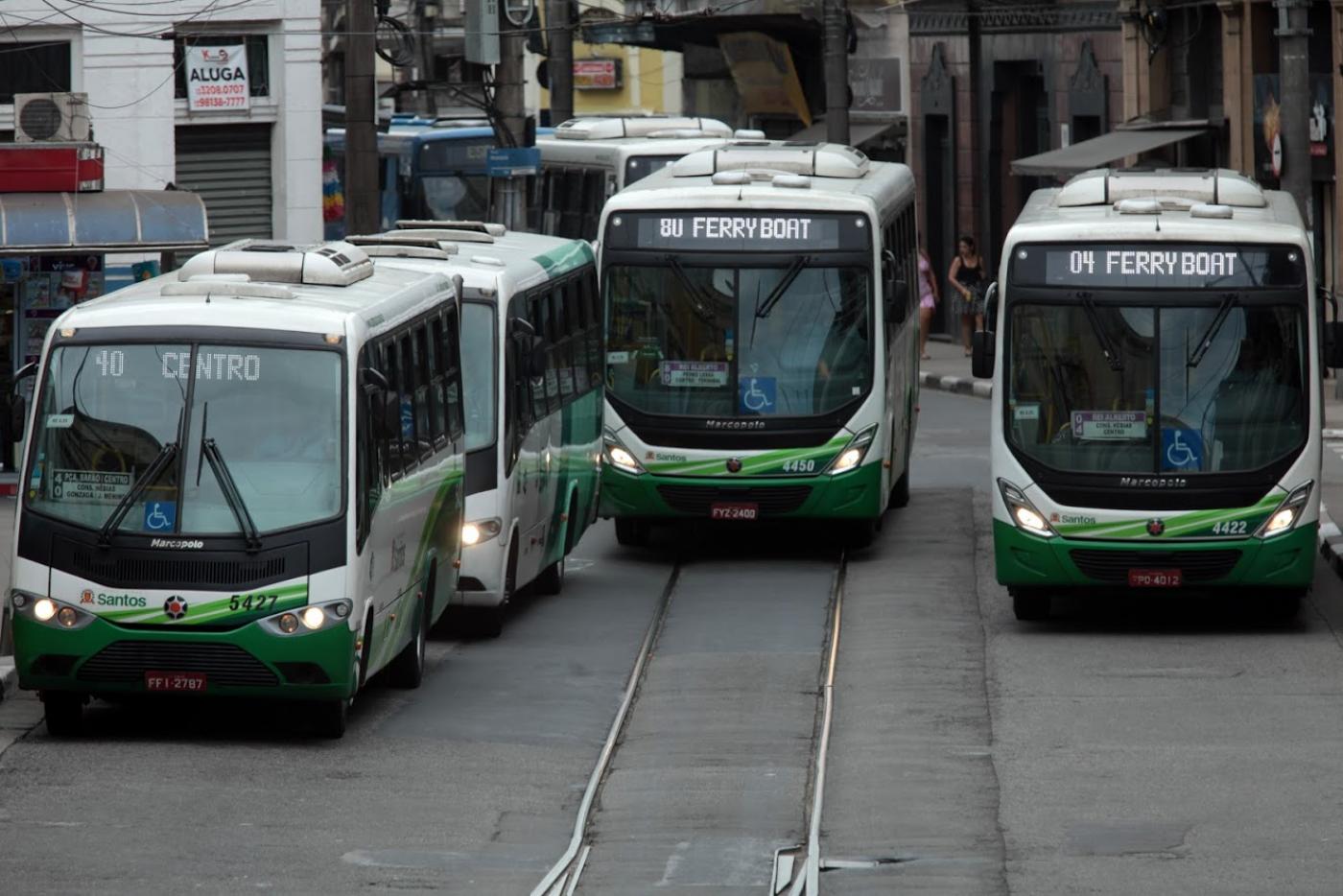 Seis ônibus em uma rua. Eles fazem fila à esquerda e outros dois estão passando ao lado. #Pracegover 