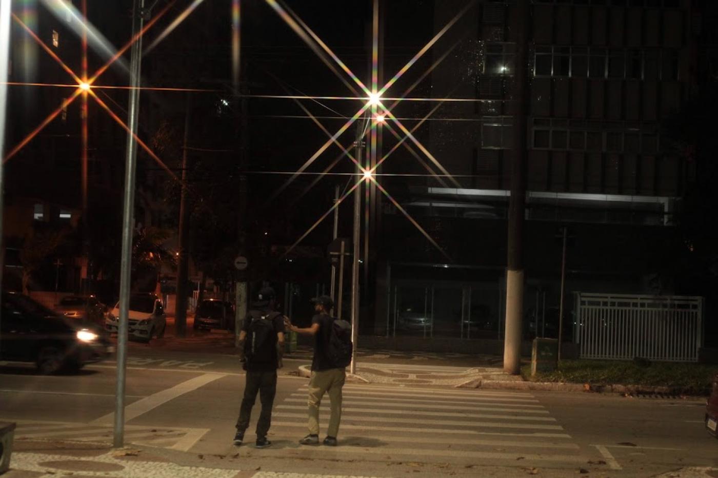 duas pessoas estão atravessando faixa iluminada à noite. #paratodosverem 