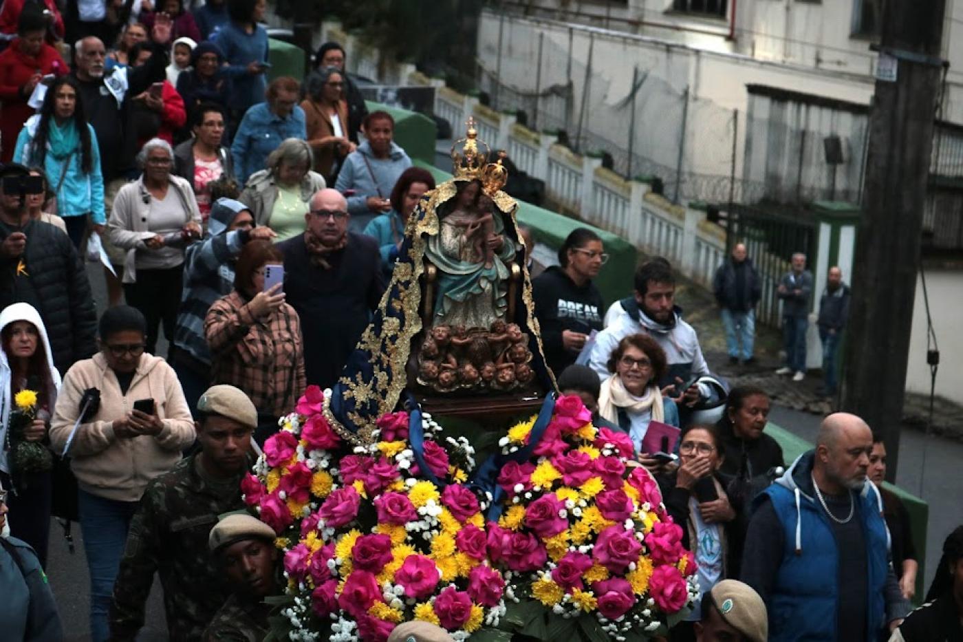 imagem de santa é carregada sobre andor e ladeada por flores. Fieis seguem a imagem que desce o morro. #paratodosverem 