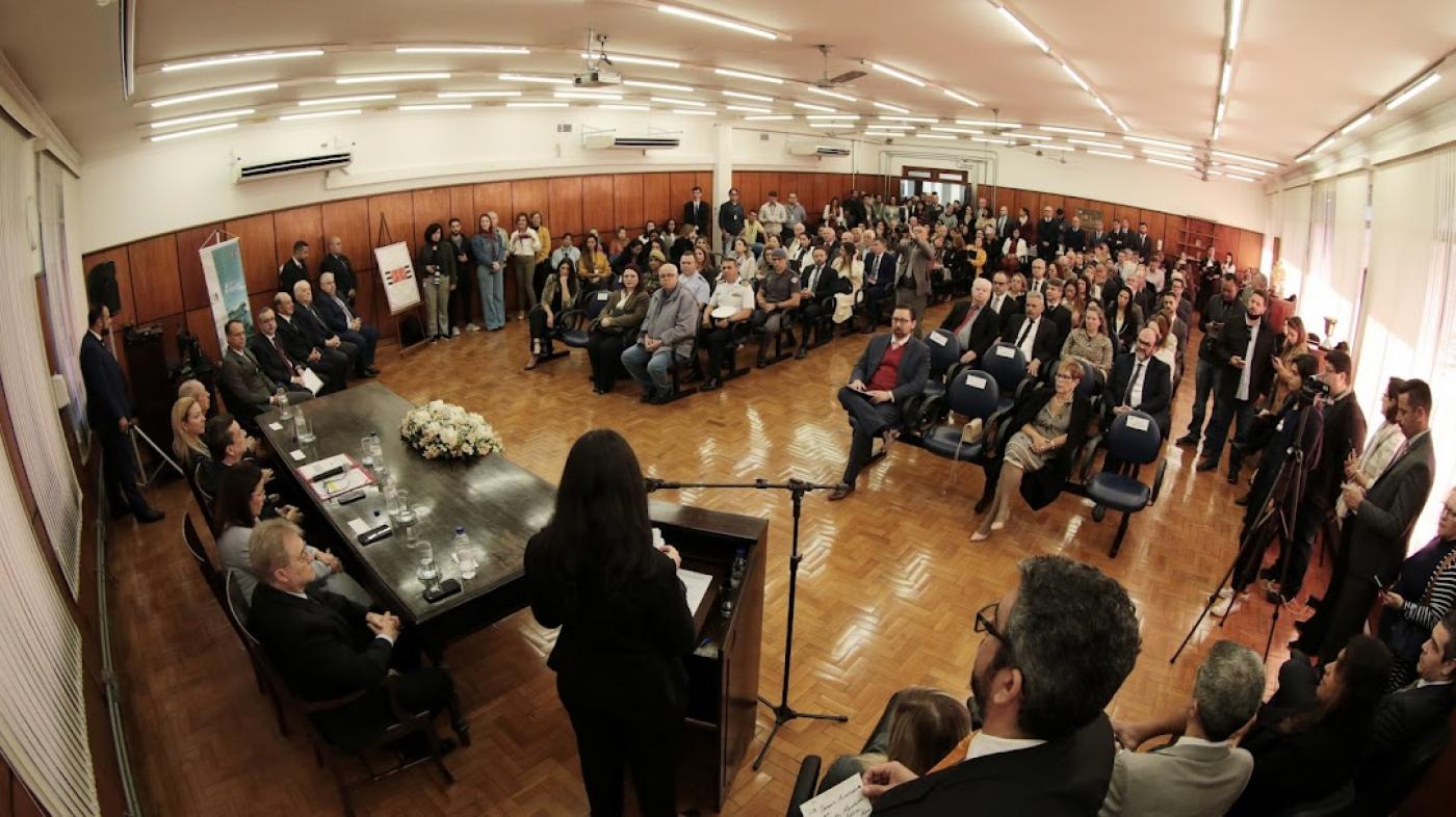 salão com várias pessoas sentadas e mesa com autoridades à frente do público. #paratodosverem 