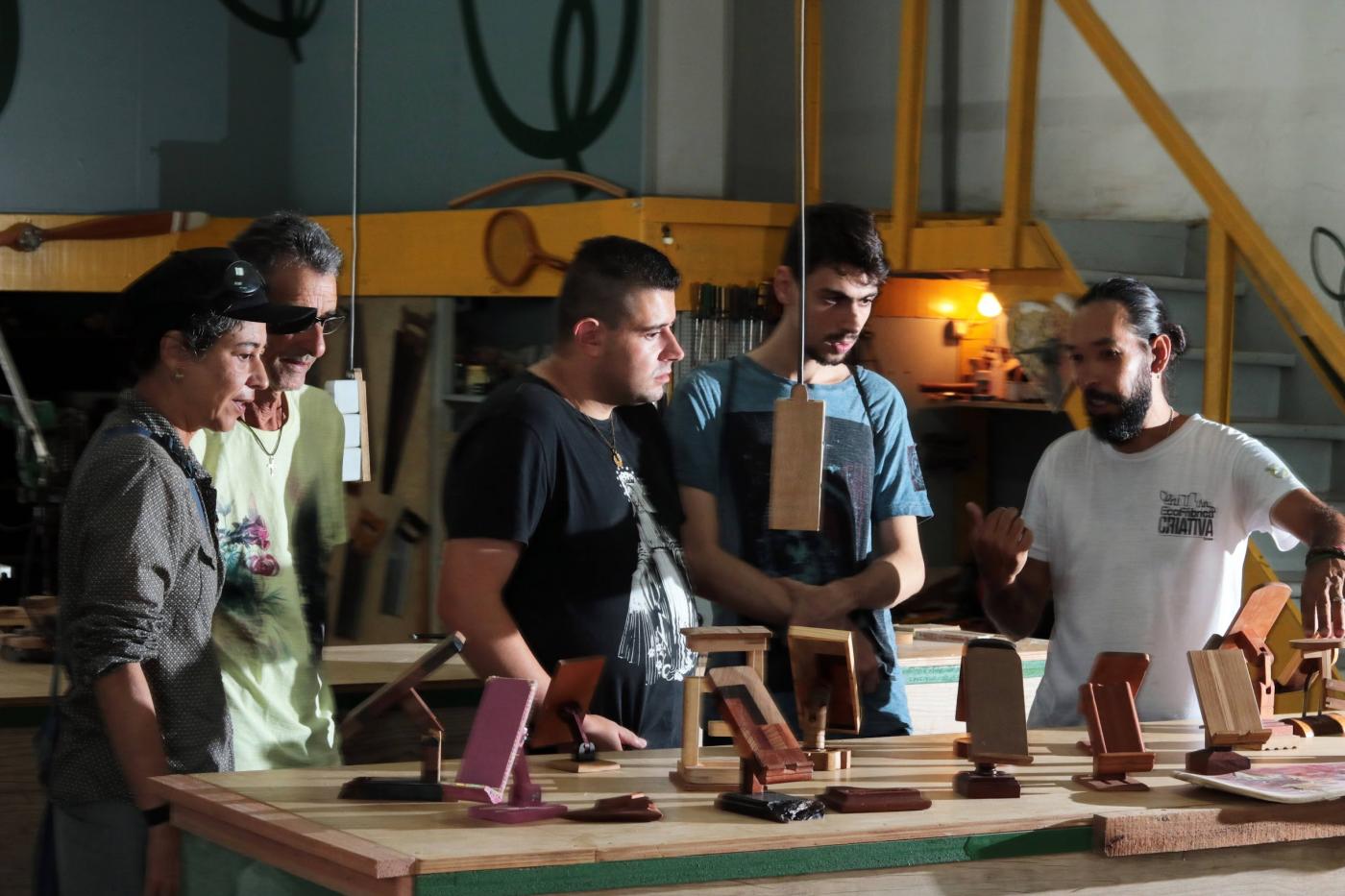 cinco pessoas estão em pé diante de uma mesa com peças em madeira. um deles, o professor fala ao grupo