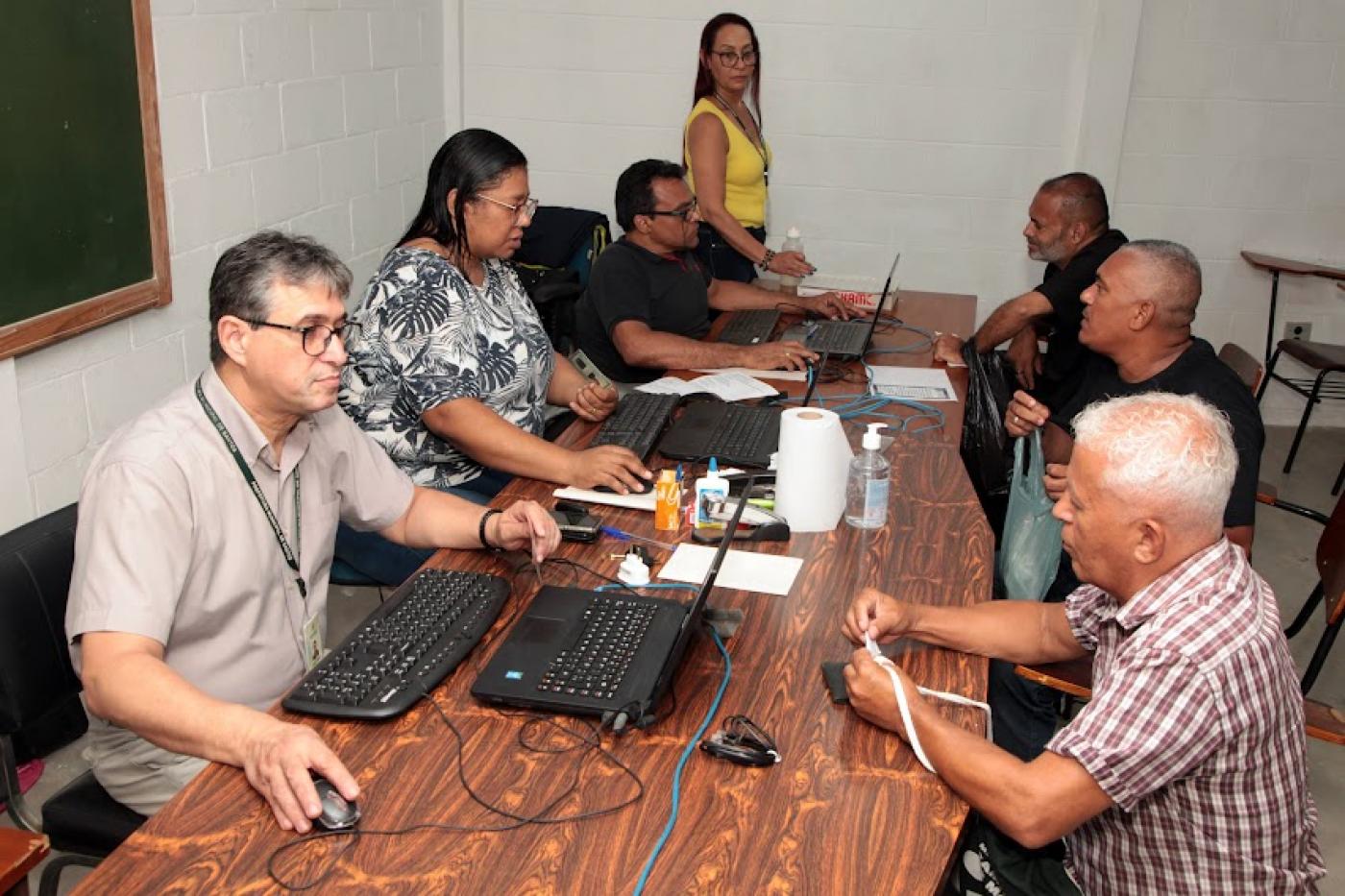 pessoas estão sentadas em torno de mesas. De um lado, o pessoal de atendimento, com computadores abertos. Do outro, pessoas fazendo consultas. #paratodosverem