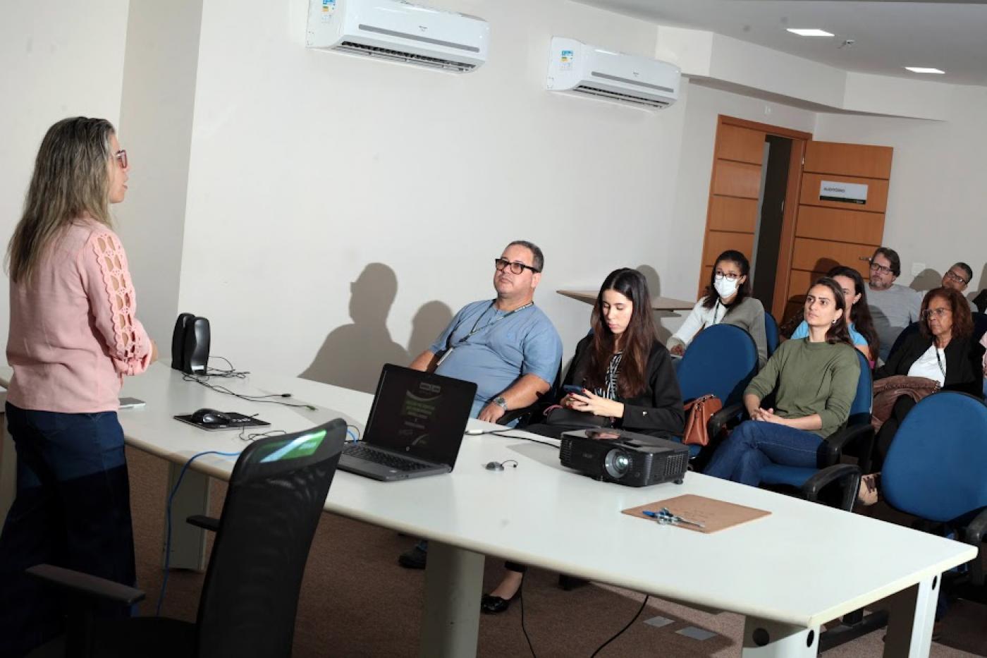 pessoas sentadas em auditório assistindo palestra. Uma mulher está em pé atrás de uma mesa falando a todos. #paratodosverem