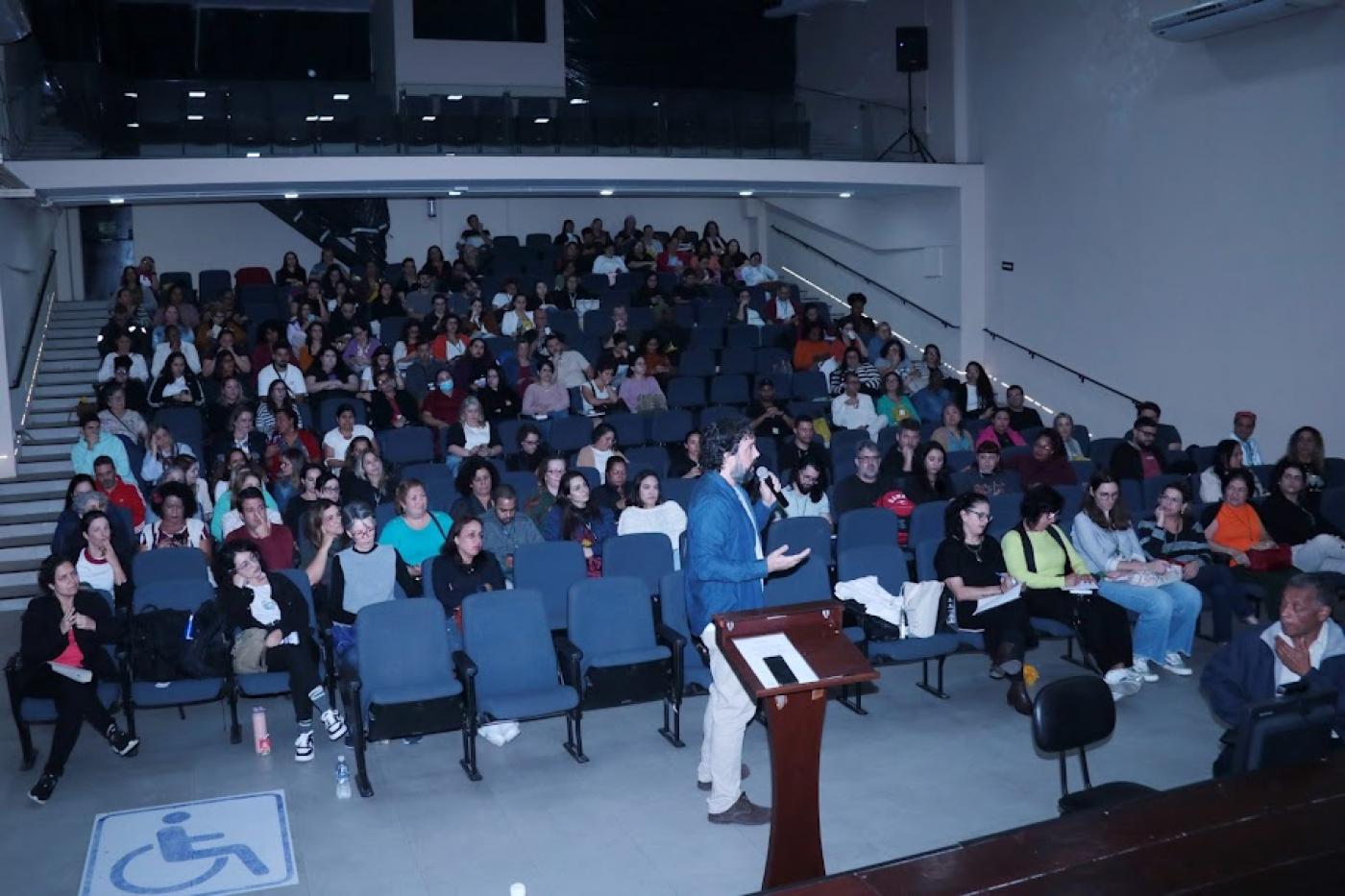 público está sentado em um auditório assistindo palestra de um homem que fala ao microfone à frente de todos. #paratodosverem 