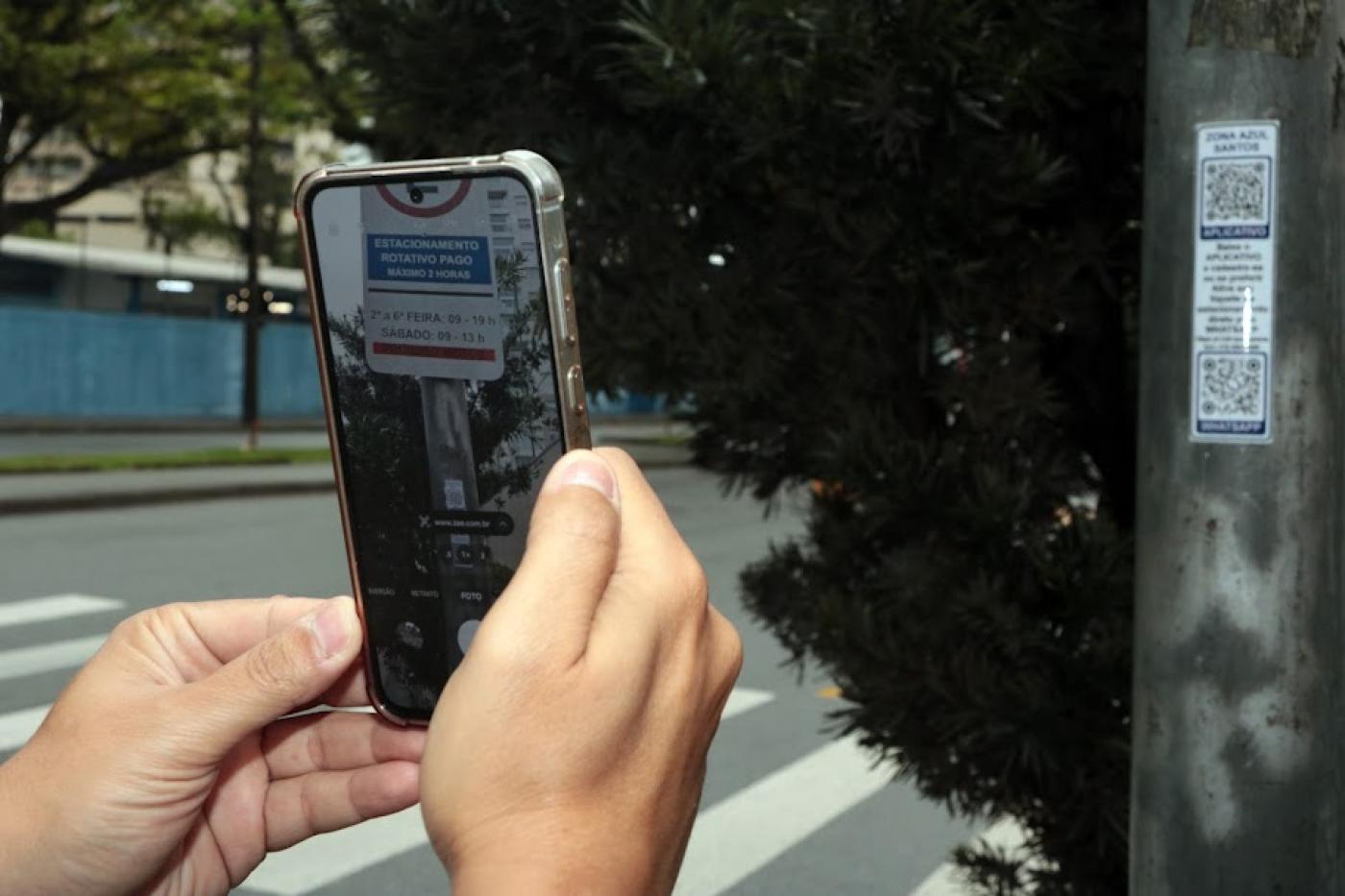 mãos seguram celular em cjua tela se fotografa o adesivo e aparece a placa da zona azul afixada em um poste que aparece à direita. #paratodosverem 