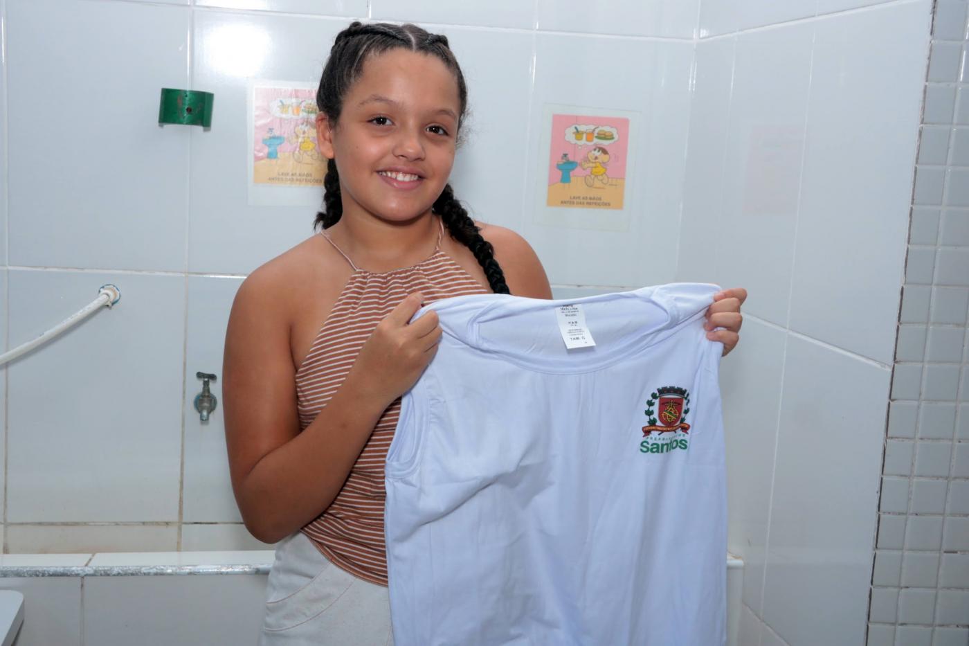 menina sorri para foto segurando camiseta de uniforme aberta pelas duas mãos. #paratodosverem 