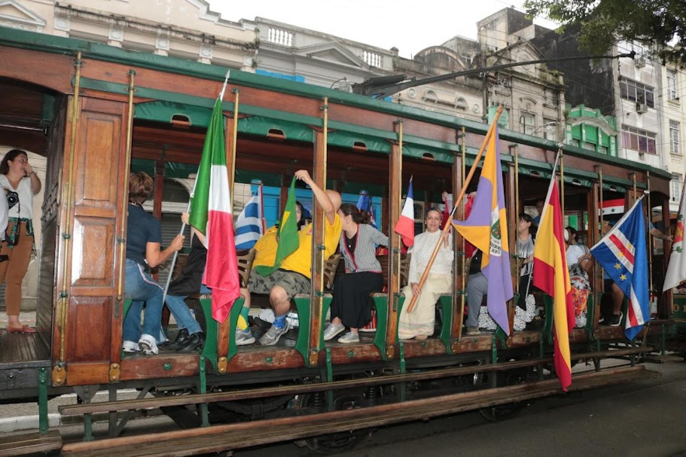 bonde com pessoas carregando bandeiras de vários países por fora do carro. #paratodosverem 