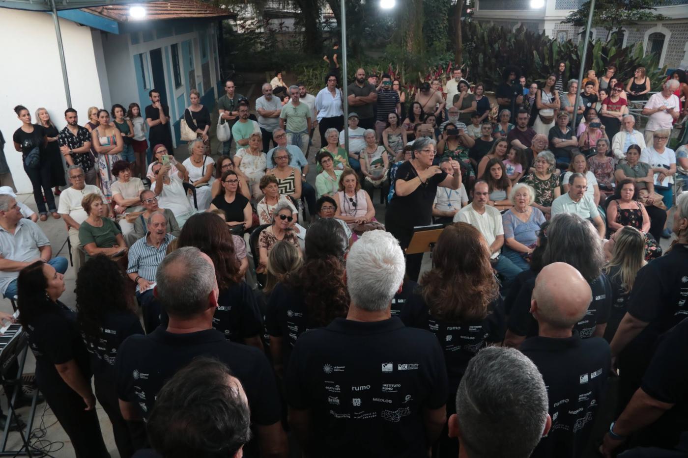 mulher rege coral diante de plateia ao fundo. #paratodosverem