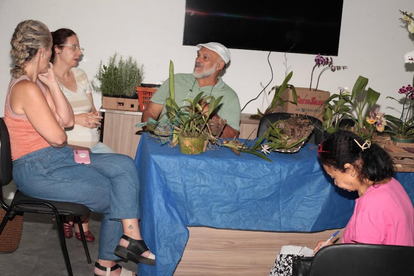 homem etá sentado atrás de uma mesa onde há várias mudas de plantas. Duas mulheres sentadas ao lado conversam com ele. #paratodosverem 