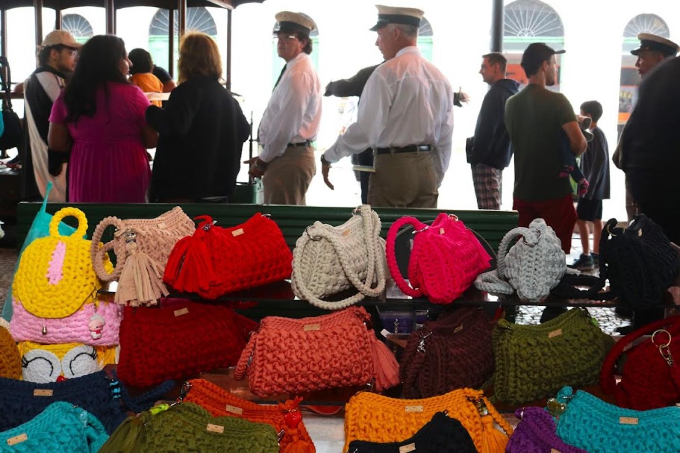 bolsas de crochê sobre mesa de exposição con pessoas circulando ao fundo. #paratodosverem 