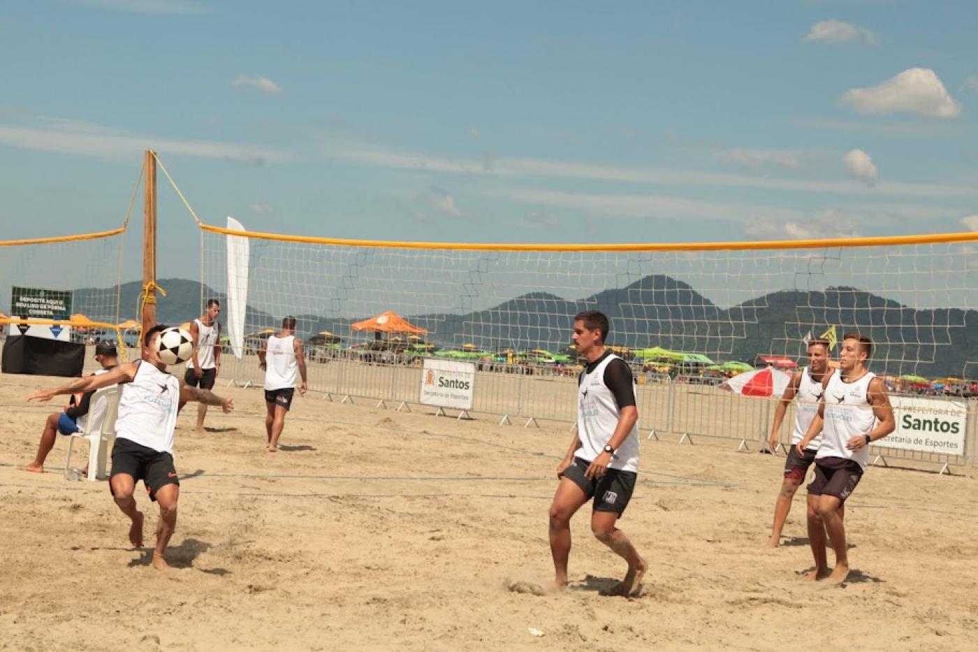 jovens estão jogando futevôlei na areia. Um deles aparece com a bola à frente do rosto, próximo ao peito, em movimento. Outros estão atentos à bola. #paratodosverem 