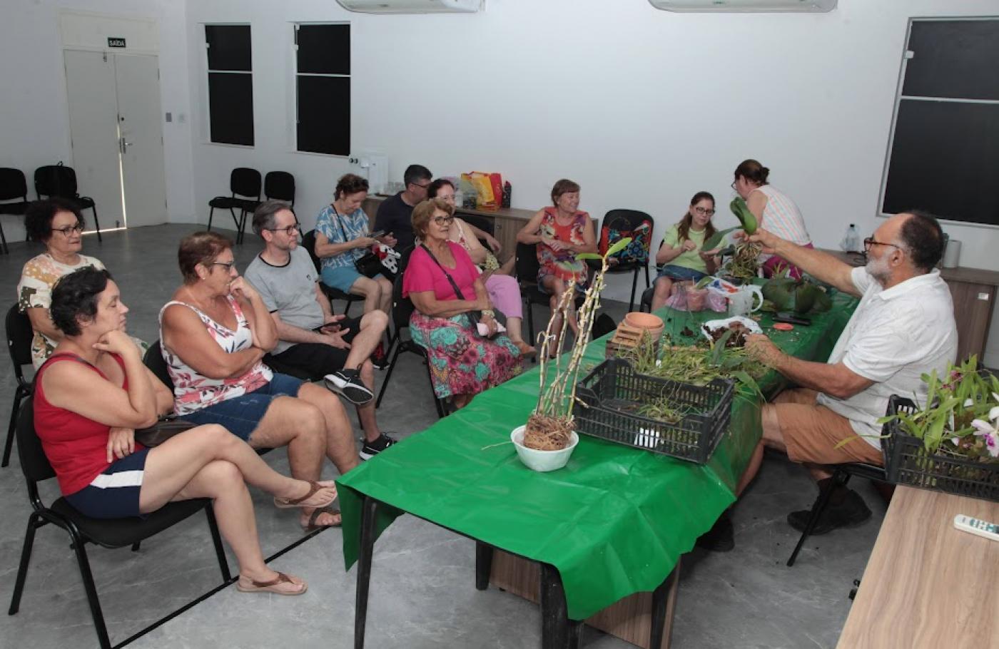 pessoas estão sentadas em auditório assistindo a um momente que fala sentado diante de uma mesa sobre a qual há vários vasos de orquídeas. #paratodosverem 