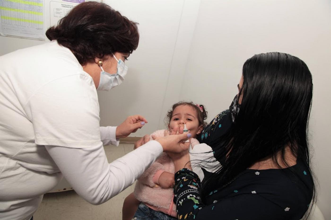 mulher vacina com a gotinha uma criança que está no colo da mãe. #paratodosverem 