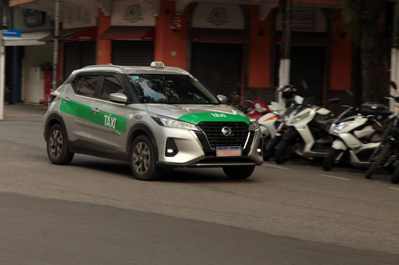 táxi passando por uma rua. Atrás há motos estacionadas junto ao meio-fio. #paratodosverem 