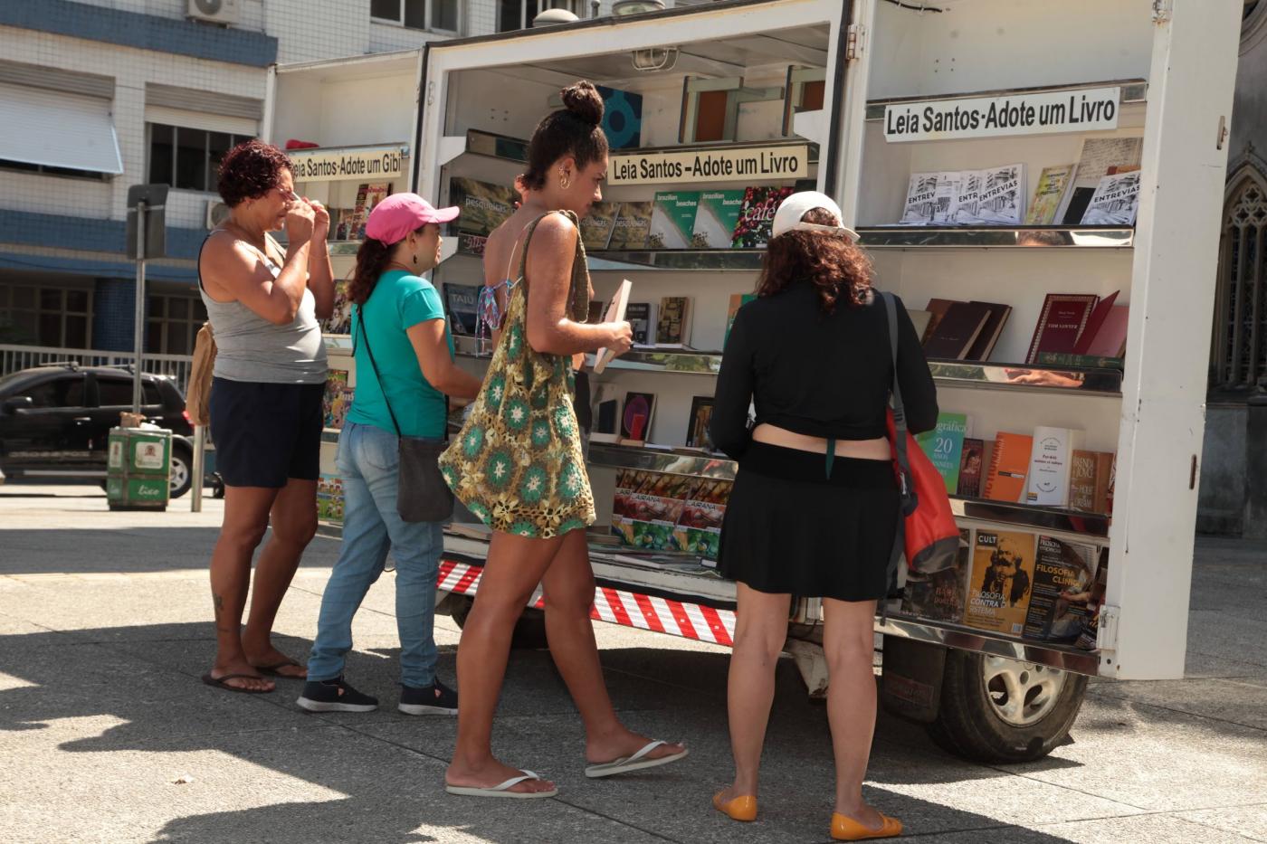 estande com livros expostos em praça. Pessoas manuseiam exemplares. #paratodosverem 