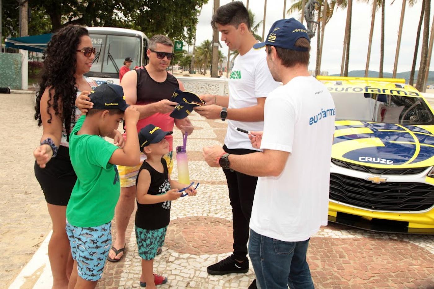 veículo da stock car ao fundo e pilotos conversando com o público à frente. #paratodosverem