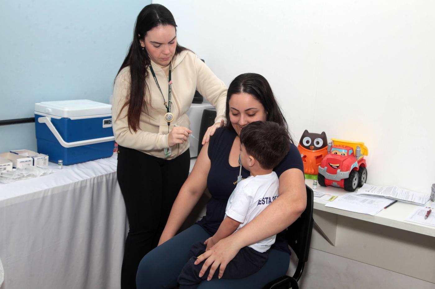 mulher aplica vacina em criança no colo de mãe. #paratodosverem