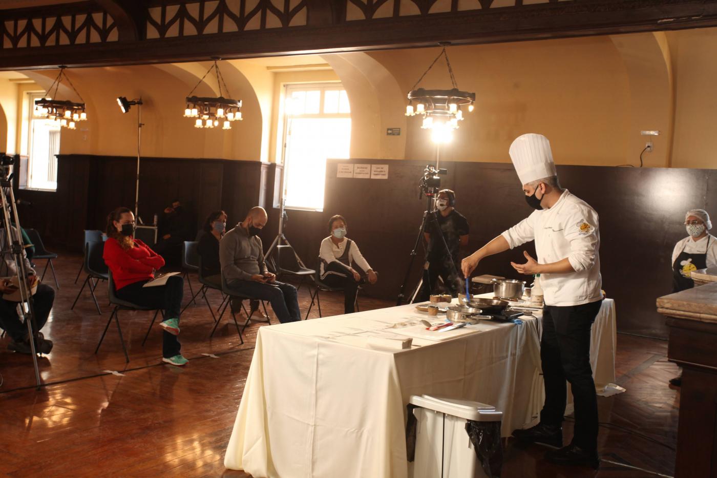 #pracegover Com uniforme de chef de cozinha, Júnior Monteiro maneja talheres sobre mesa à frente de câmeras 